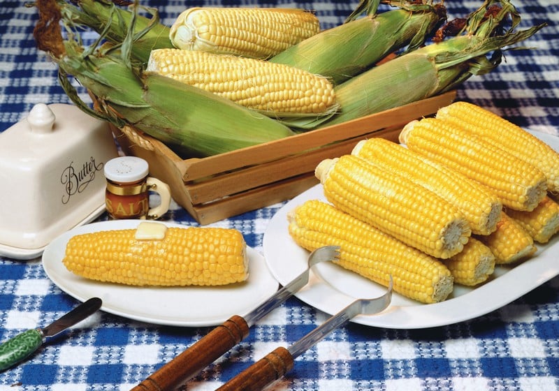 Ears of corn on white plates, in wooden baskets, with butter on navy cloth table Food Picture
